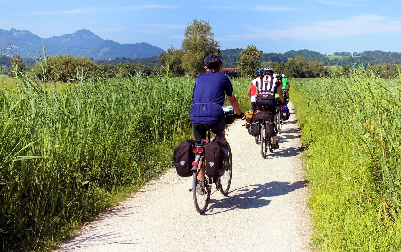 Radfahrer mit Fahrradtaschen auf Radweg zwischen grünen Feldern. Im Hintergrund Berge