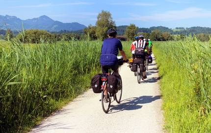 Radfahrer mit Fahrradtaschen auf Radweg zwischen grünen Feldern. Im Hintergrund Berge